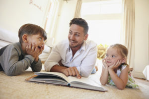Shot of happy young family lying on the floor reading a book. Young man reading stories to his little son and daughter at home.